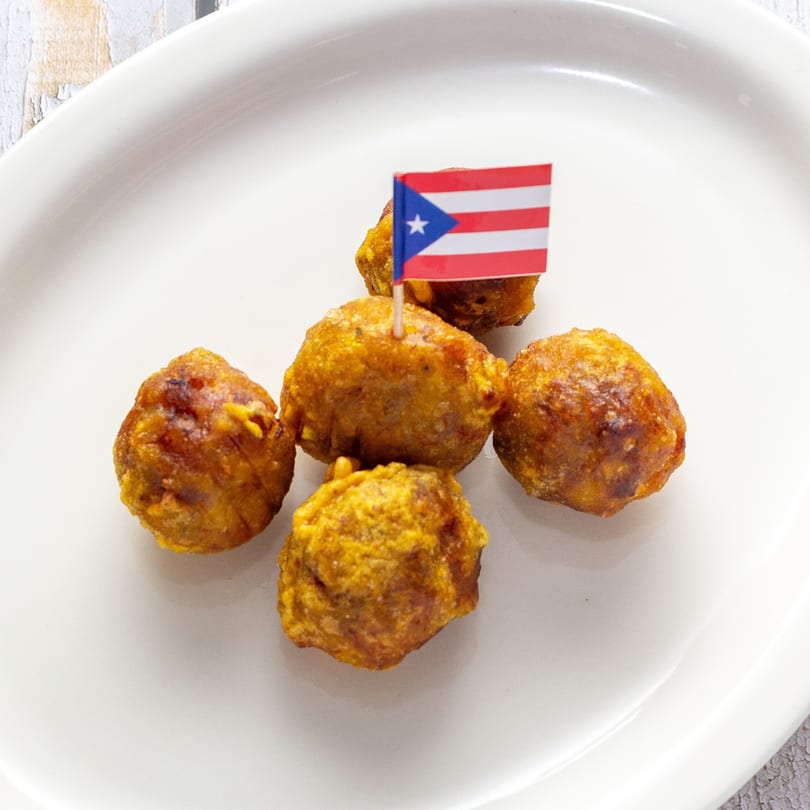 Five golden-brown Puerto Rican fritters, one topped with a small Puerto Rican flag, on a white plate.