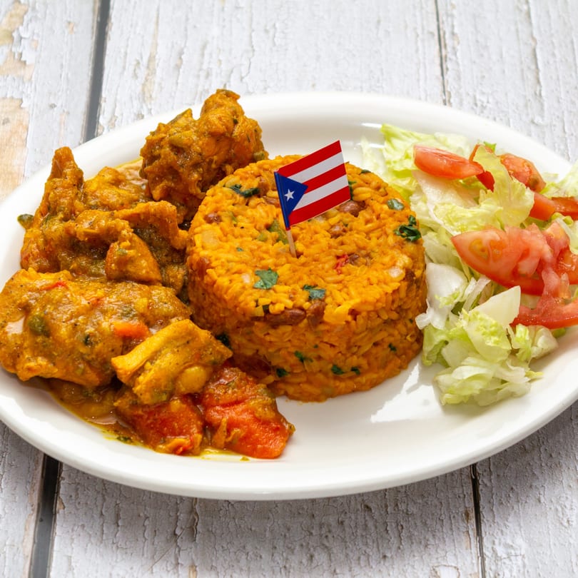 Puerto Rican chicken stew with yellow rice, a small flag, and a side salad on a white plate.