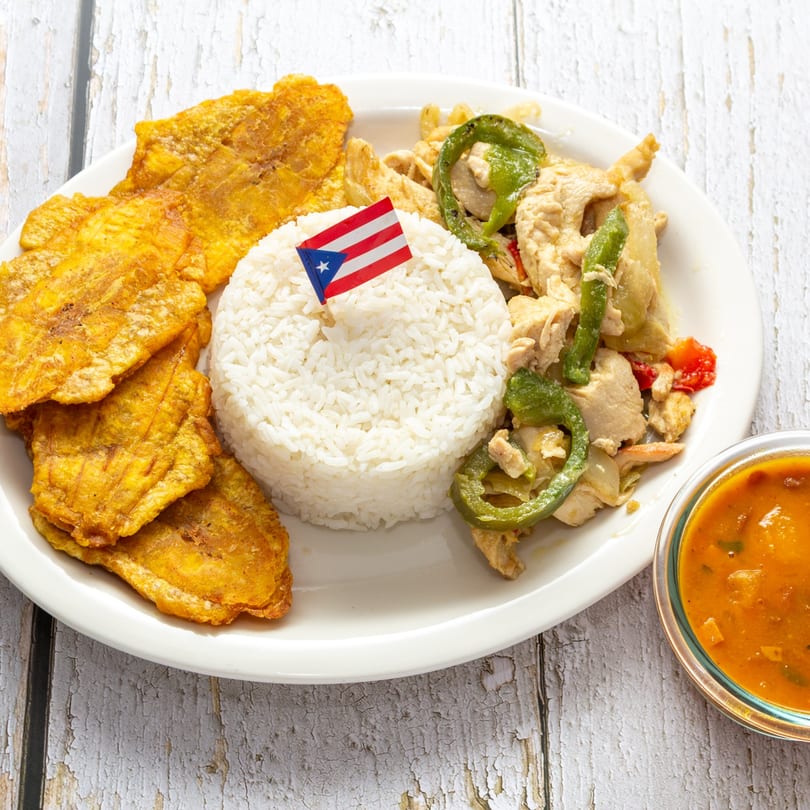 Puerto Rican chicken with peppers, white rice, fried plantains, and a side of sauce.